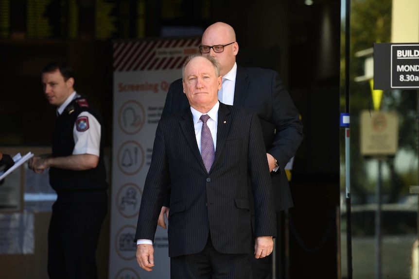 A man in a suit leaves court with security in the background.