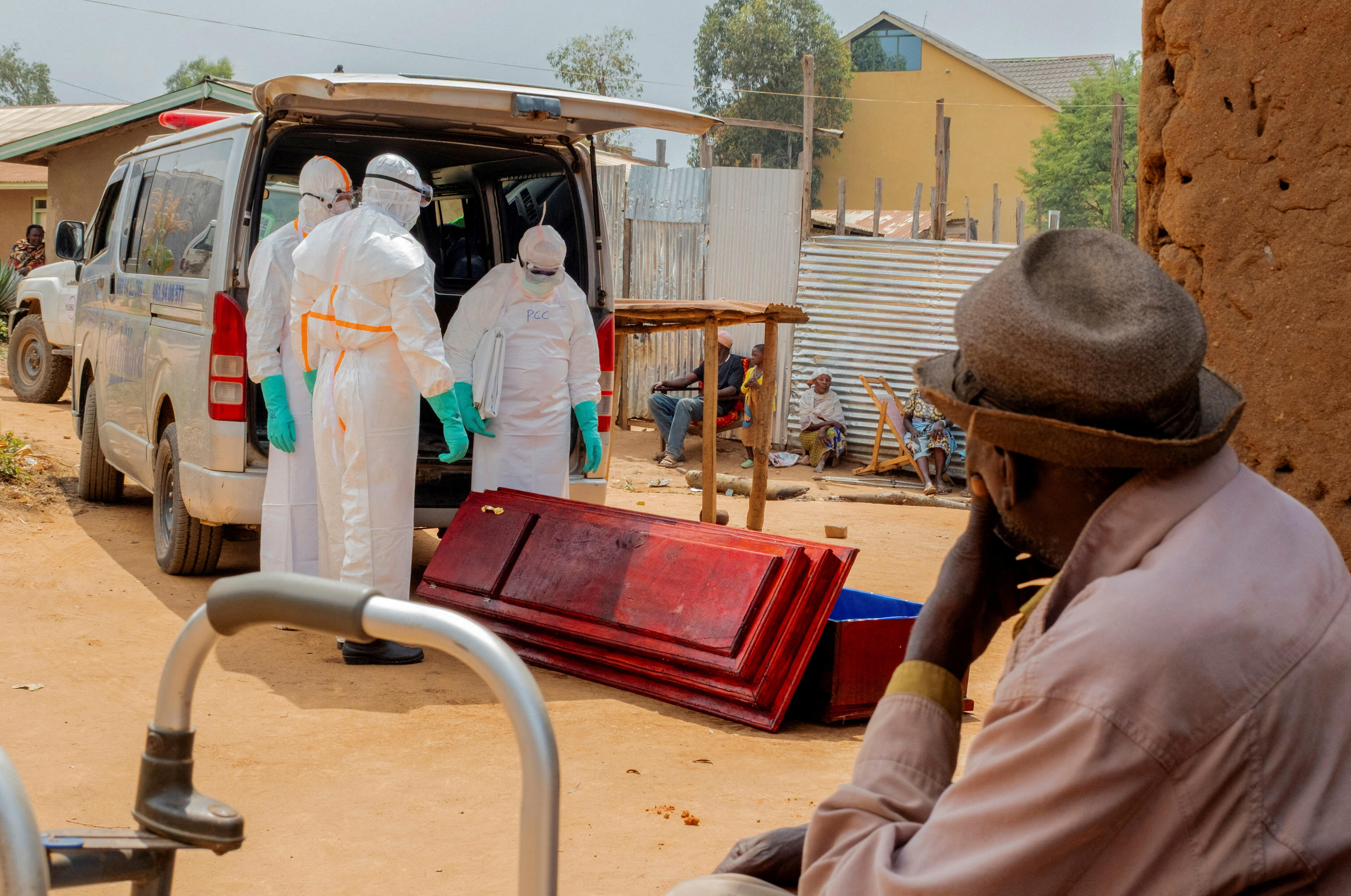 A man wearing a hat sits by as three men in full body, white personal protective equipment stand around a coffin.