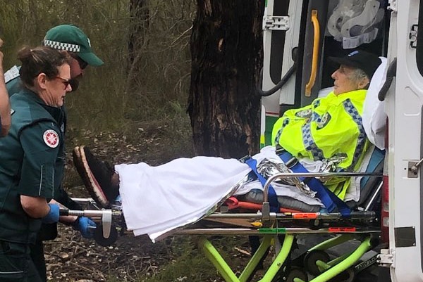 A woman in a hi-vis WA Police jacket lies on a gurney as two St John Ambulance personnel load it into the back of an ambulance.