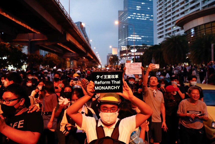 A group of people wearing masks and holding up a sign saying Reform the Monarchy march in a street.