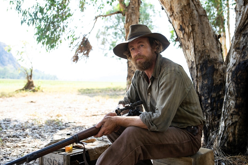 Simon Baker sits on a log, in his role as ex-soldier Travis in the 2020 film High Ground.