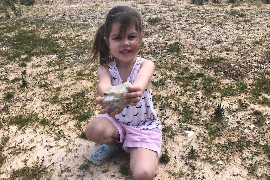 A smiling toddler with hair tied on one side, wears pink top and shorts, kneals on ground, holds out a large stone.