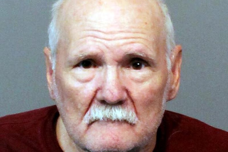 An older man mostly bald with grey sideburns and white moustache looks vacantly during police mugshot 
