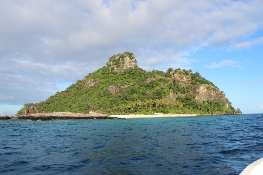 Blue ocean surrounds a deserted small island.  Large rock face and palm trees, green shrub visible from sea.