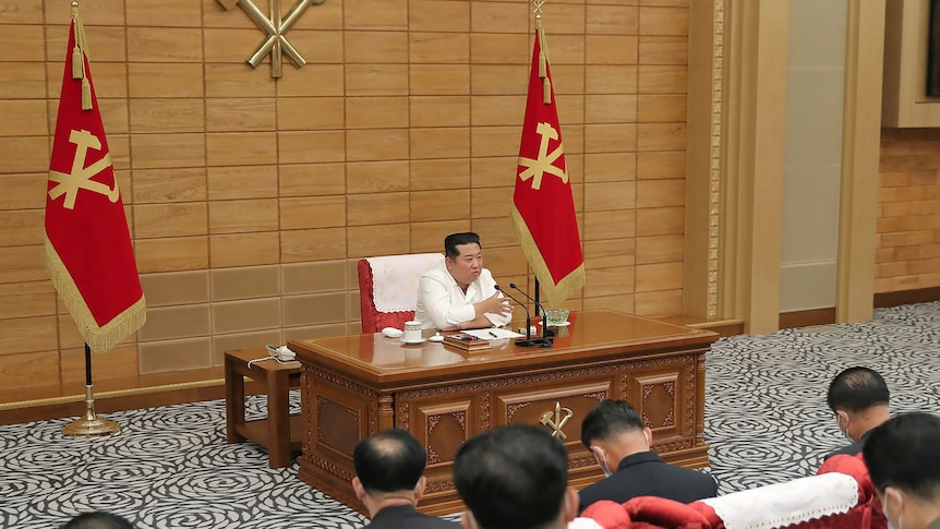 Kim Jong Un in a white suit sits at a desk in a room with two red flags either side of him as men in suits sit in the audience