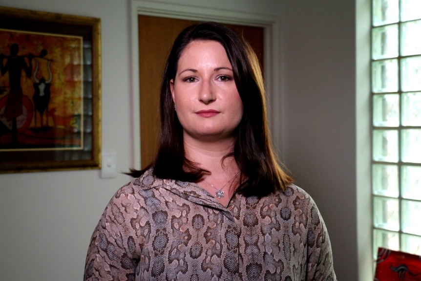 A woman wearing a printed long-sleeved top stands next to a painting and in front of a door.