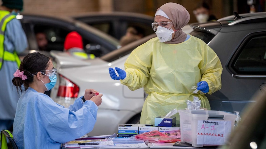 two women in scrubs and PPE holding test tubes surrounded by cars