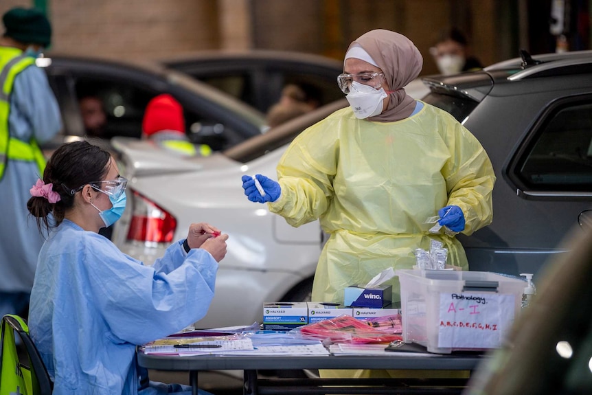 two women in scrubs and PPE holding test tubes surrounded by cars