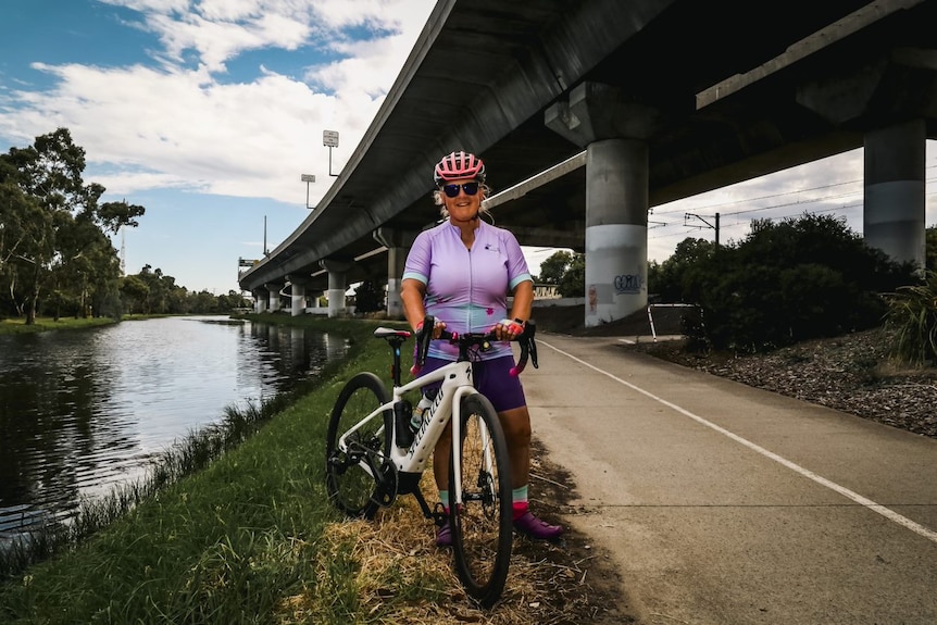 Middle aged woman dressed in mauve lycra poses with her bike beside a designated bike path