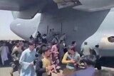 a large group of people crowd around a grey military transport plane, some grabbing on to the side of it.