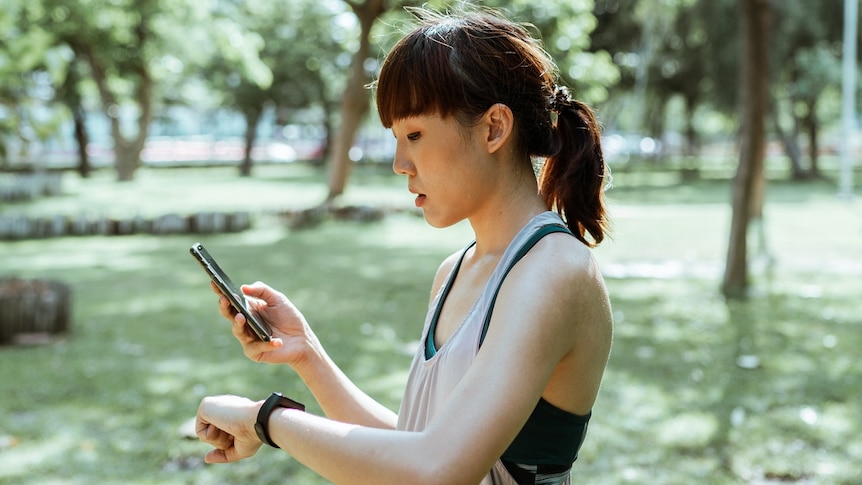 A woman in athleisure wear stands in a park, holding her smartphone in one hand while looking at her fitness tracker on her arm
