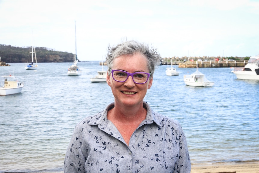 A middle-aged woman withy grey hair and purple glasses smiles with boats in a bay behind her