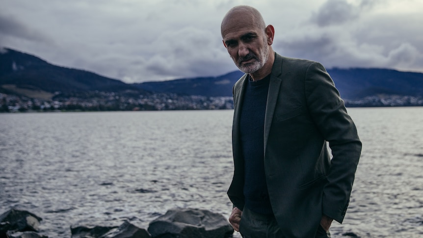 Paul Kelly stands on the bank of a body of water, looking unsmiling at the camera. He wears a jacket and dark shirt.