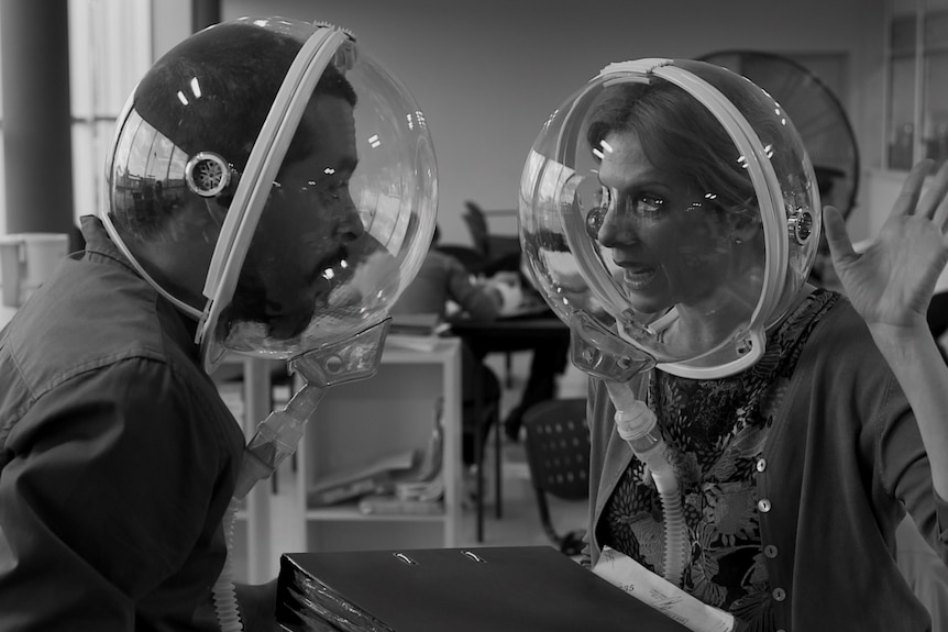 A black and white shot of a man and a woman in conversation in an office, wearing astronaut-like bubbles on their heads.
