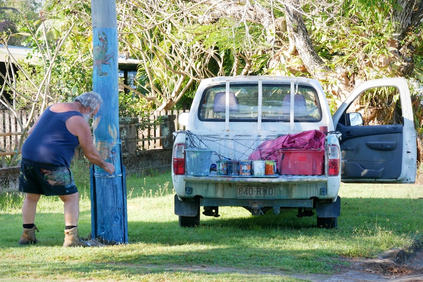 A man paints a power pole next to a ute tray filled with paint cans