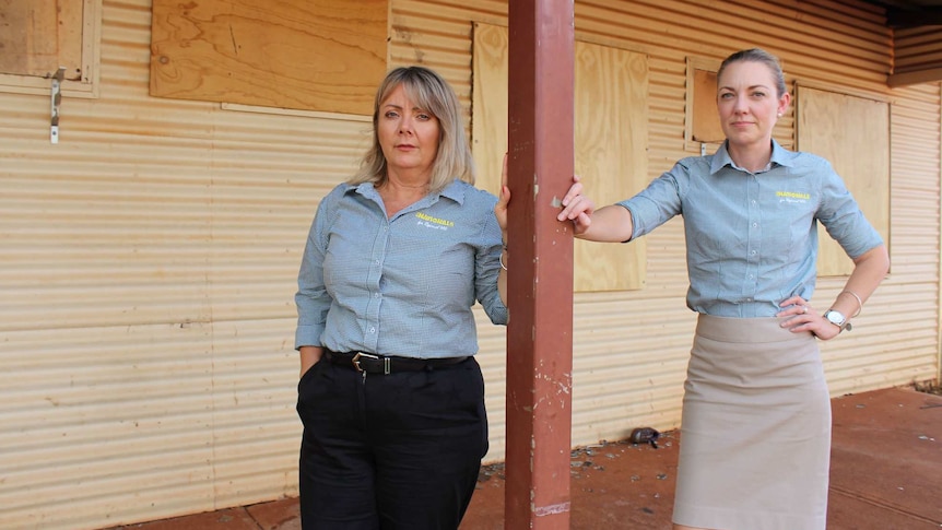 A wide shot of Mia Davies and Jacqui Boydell standing outside a boarded up building leaning against a pole.