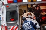 A patient on a stretcher with a mask being unloaded from an ambulance with a long line of ambulances in the background.