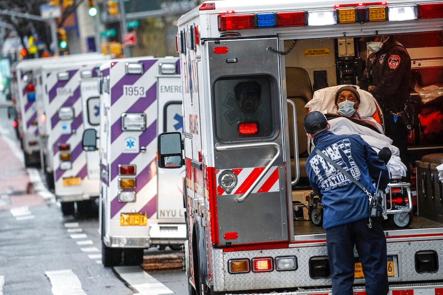 Un paciente en camilla con máscara se baja de una ambulancia con una larga fila de ambulancias al fondo.