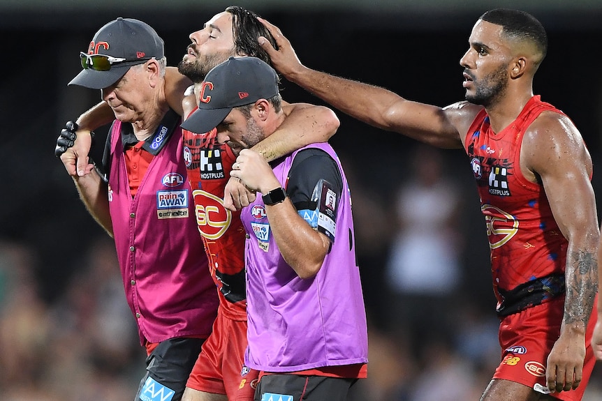 Lachie Weller is helped off the field by trainers as Touk Miller pats him on the head