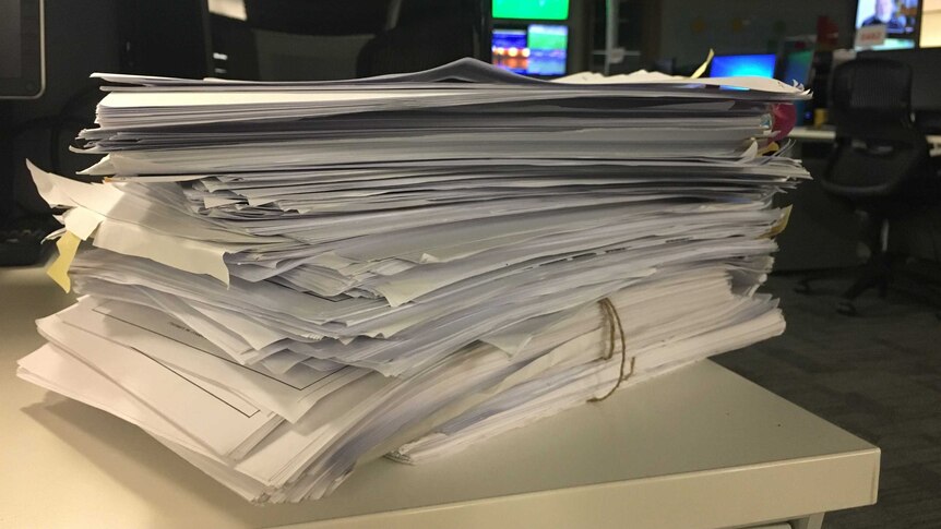 A stack of cabinet files sitting on a desk. Computer screens and TVs are visible in the background.