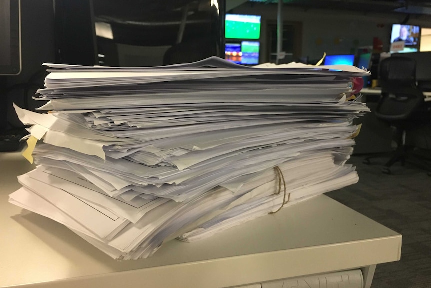 A stack of cabinet files sitting on a desk. Computer screens and TVs are visible in the background.