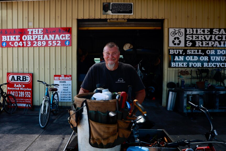 A man with a beard stands at a tool bench in front of a sign for a bike shop.