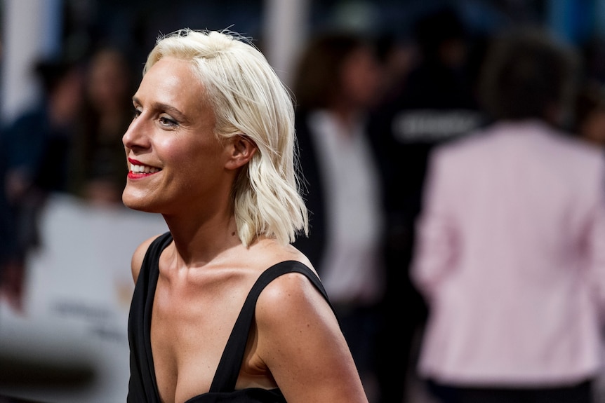 A 40-something blonde woman smiles, wearing red lipstick and a black dress, a crowd of people in the background