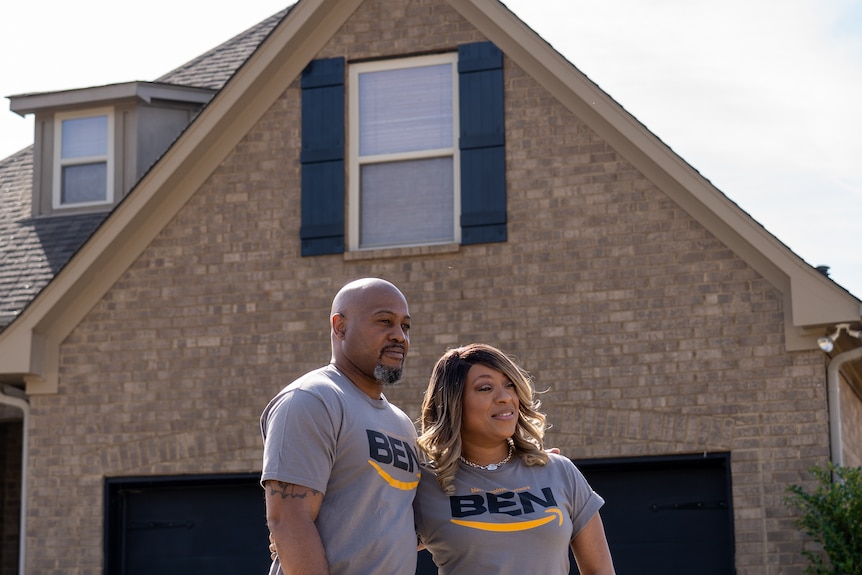 A smiling couple stand before a large brick house