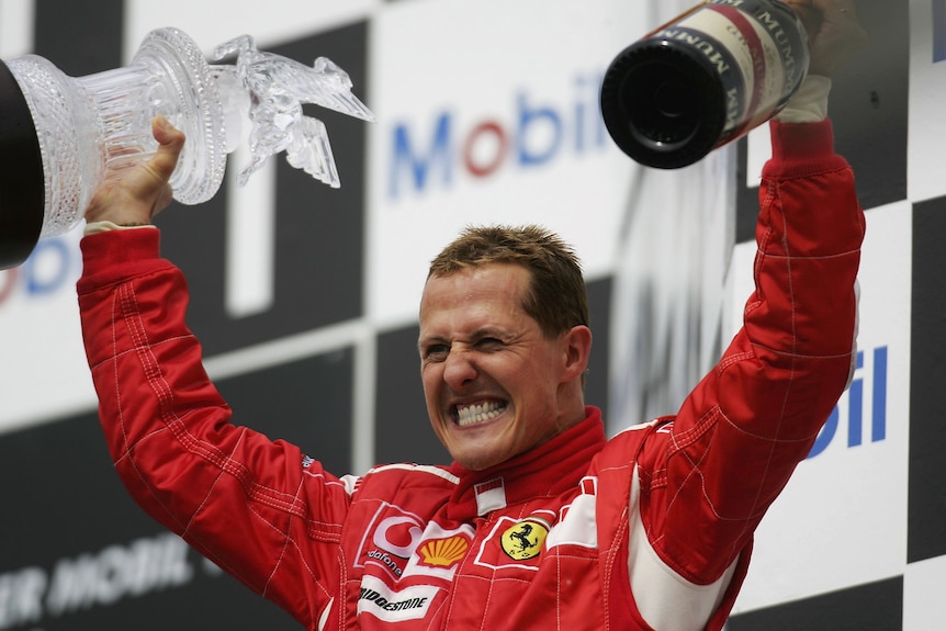 A racing driver in a red racing suit, arms raised in celebration, holding a trophy and bottle of wine.