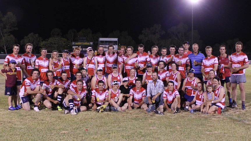 A group of men and women in red and white team shirts posing for a photograph on the Mount Isa Rugby League field.
