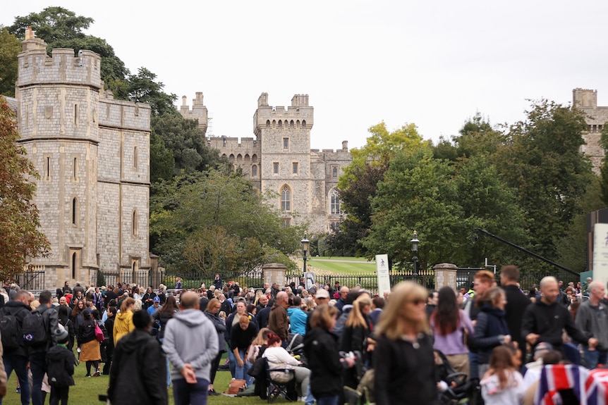 Crowds gather in a park next to a large grey castle. 