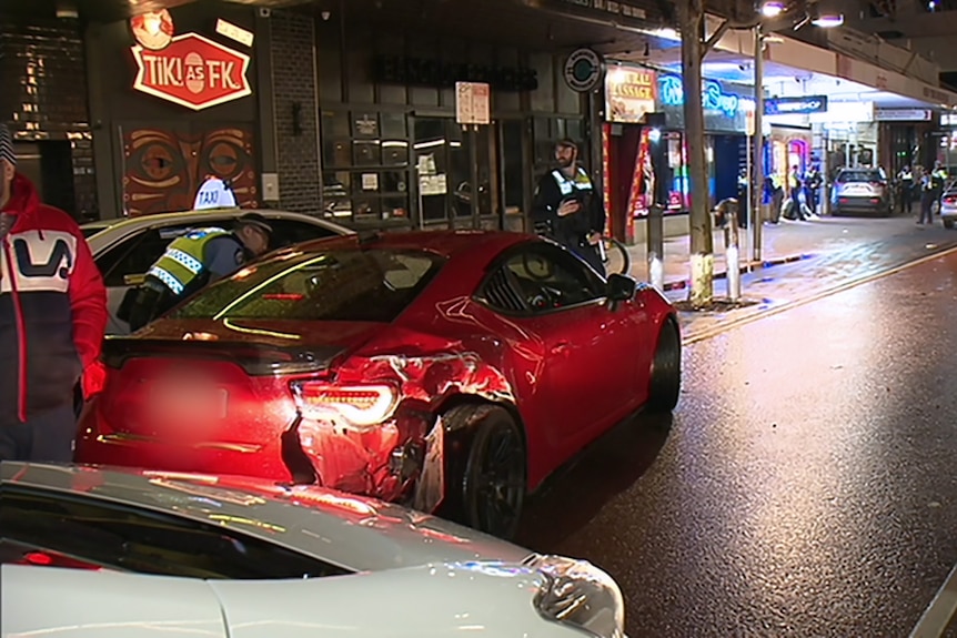 A red car with dents and crash damage in the Perth CBD at night, with police officers and pedestrians around.