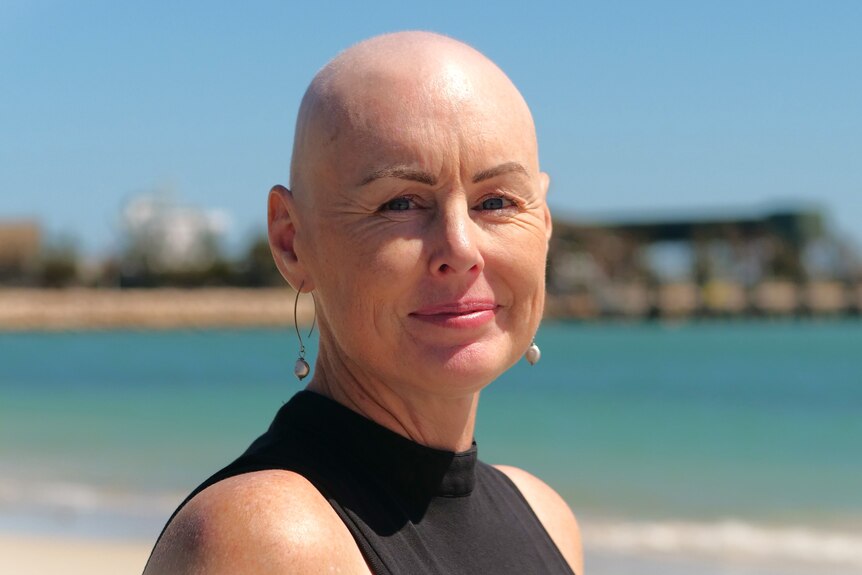 A bald woman smiles at the camera wearing a black shirt standing on the beach. 