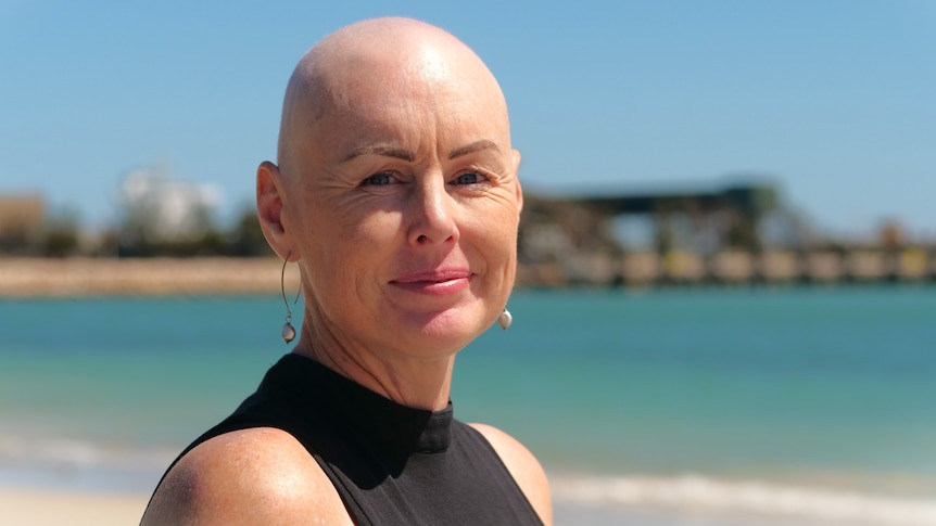 A bald woman smiles at the camera wearing a black shirt standing on the beach. 