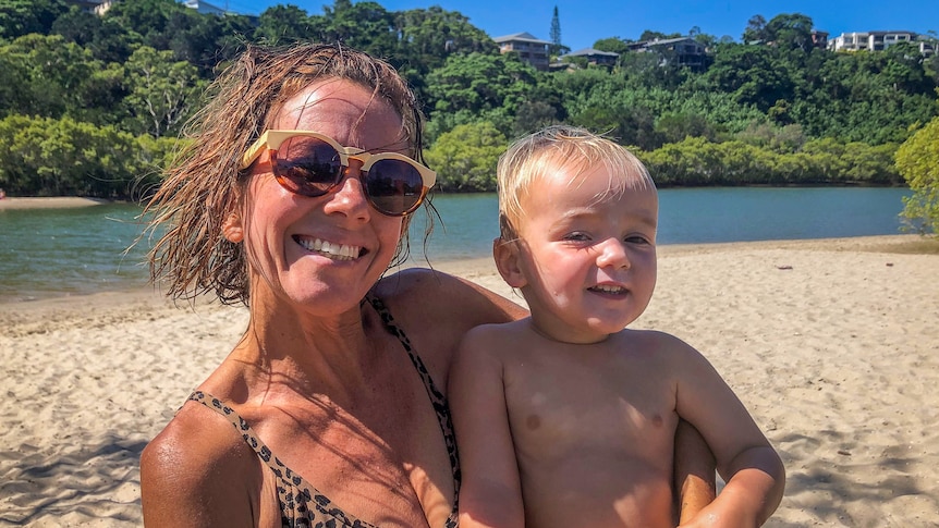 A smiling woman in bathers holds a toddler with blond hair in her arms with blue water and golden sand in the background.