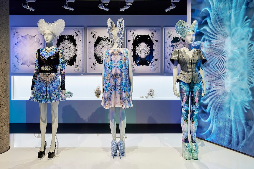Three mannequins wearing various icy blue, black and white designs stand on a platform beside a snowflake-like projection.