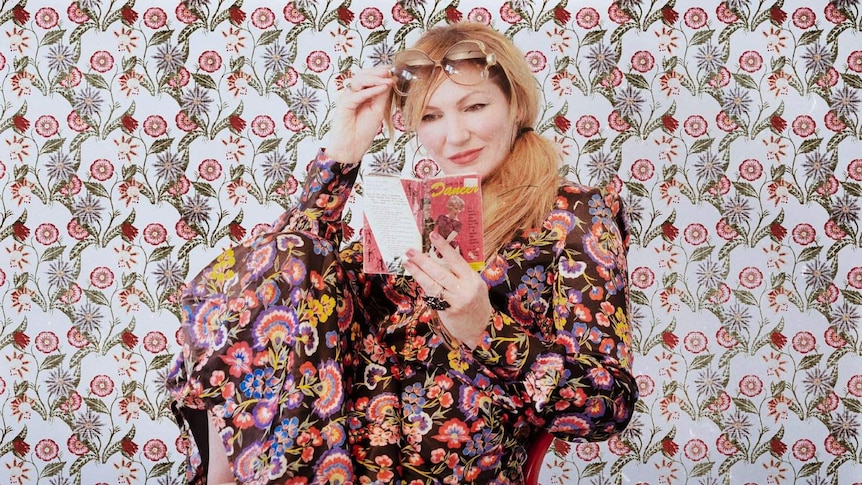 Lo Carmen sits on a chair in front of a patterned wall reading a book