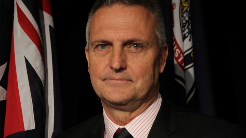 Jon Black stands in front of the Australian flag and the RSL flag. He wears an RSL pin badge on his left collar.