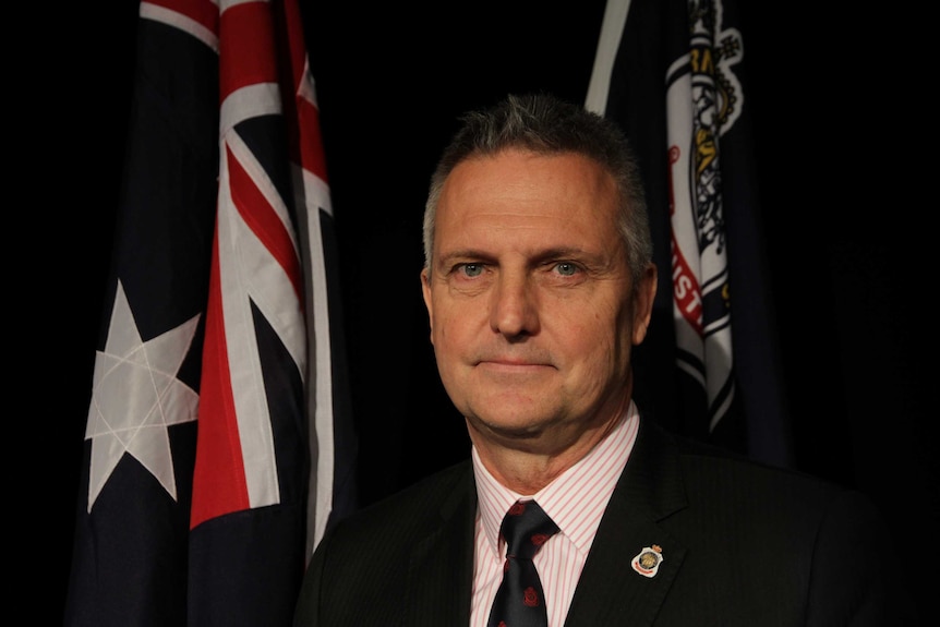 Jon Black stands in front of the Australian flag and the RSL flag. He wears an RSL pin badge on his left collar.