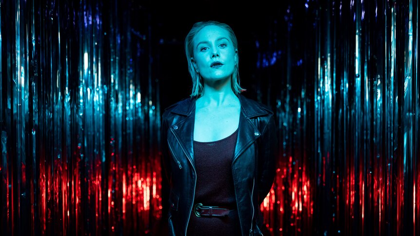 White woman with blonde lob wears black singlet and leather jacket and stand, lit in blue and red in front of silver curtain
