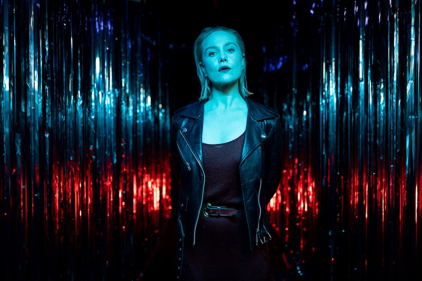 White woman with blonde lob wears black singlet and leather jacket and stand, lit in blue and red in front of silver curtain
