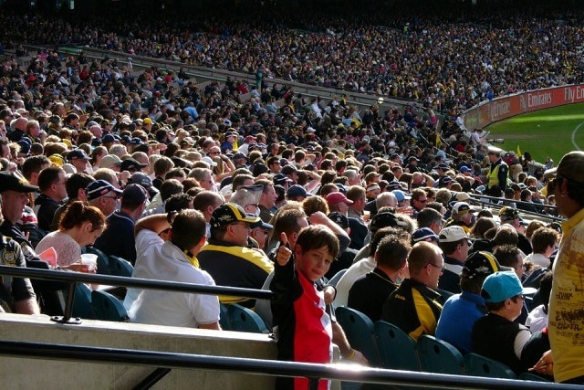 A young boy wearing an AFL team jumper stands in a packed stadium, giving a thumbs up as he turns to face the camera.