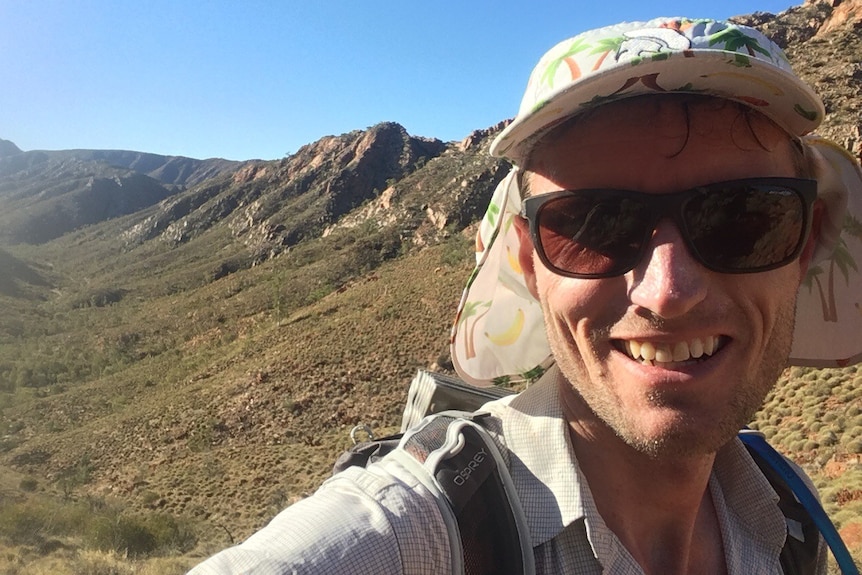 man wearing hat and sunglasses with long expanse of hilly rock behind him.