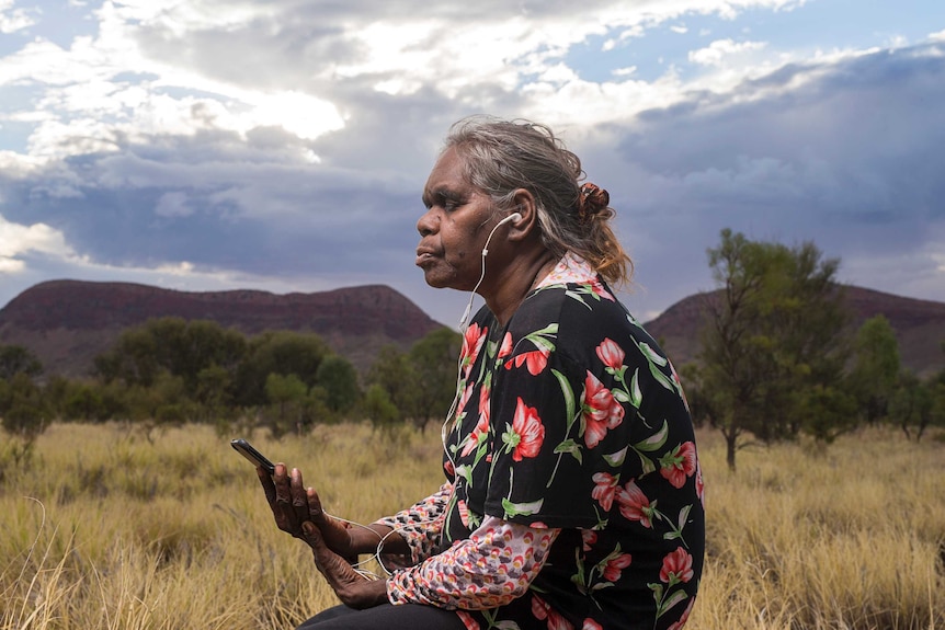 A woman holding an iPhone sits in long grass in central Australia with her eyes closed and headphones plugged in, meditating.