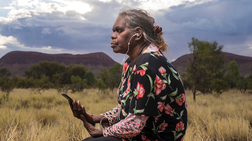 A woman holding an iPhone sits in long grass in central Australia with her eyes closed and headphones plugged in, meditating.