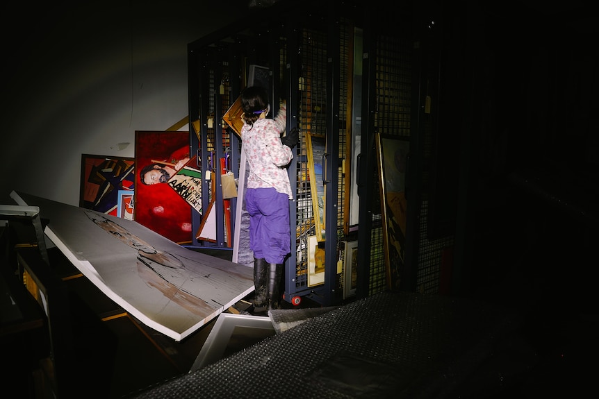 Woman with brown hair wears floral top, purple pants and gumboots opening a storage cage brimming with artworks under spotlight.