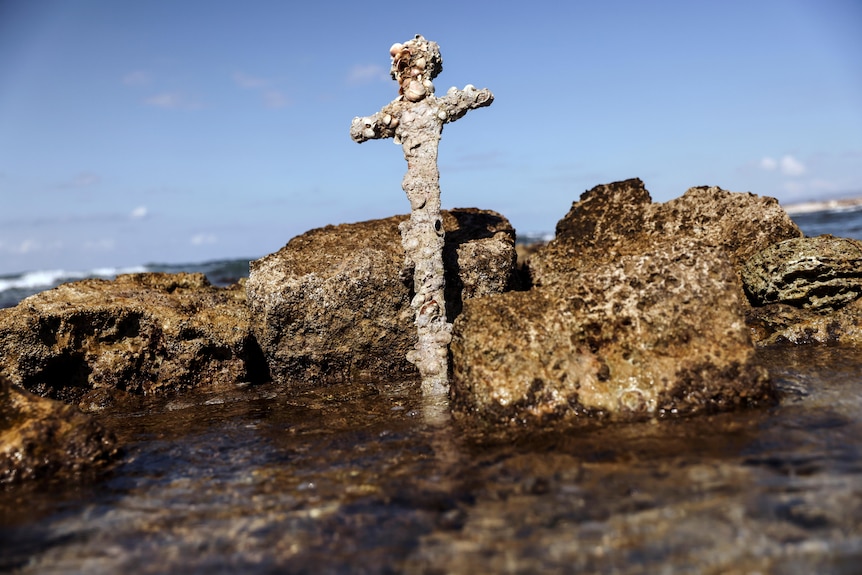A barnacle-encrusted sword leans against a rock in the shallows of a rocky sea shore.
