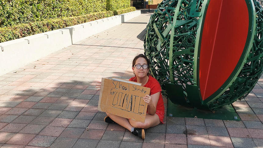 A small bespectacled girl sits on a footpath next a watermelon sculpture protesting for action on climate change.