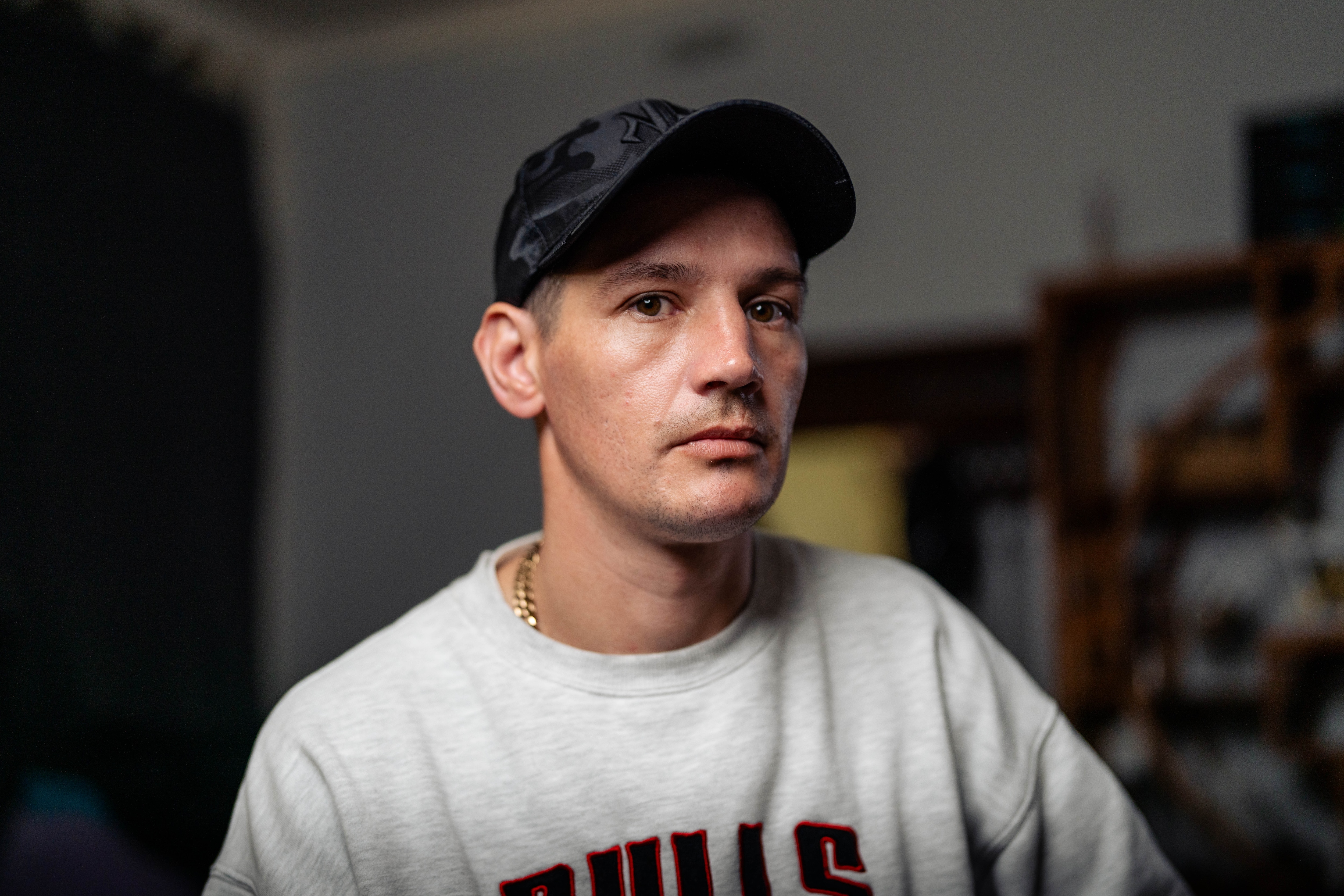 Close up of Shannon, wearing a grey jumper and a black hat, sits on a stool in a dim living room with a serious expression.