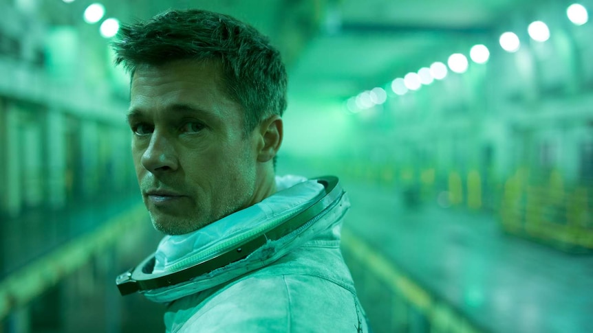 Close-up of Brad Pitt in green-lit space-type hold, wearing space suit but no helmet, looking at camera.